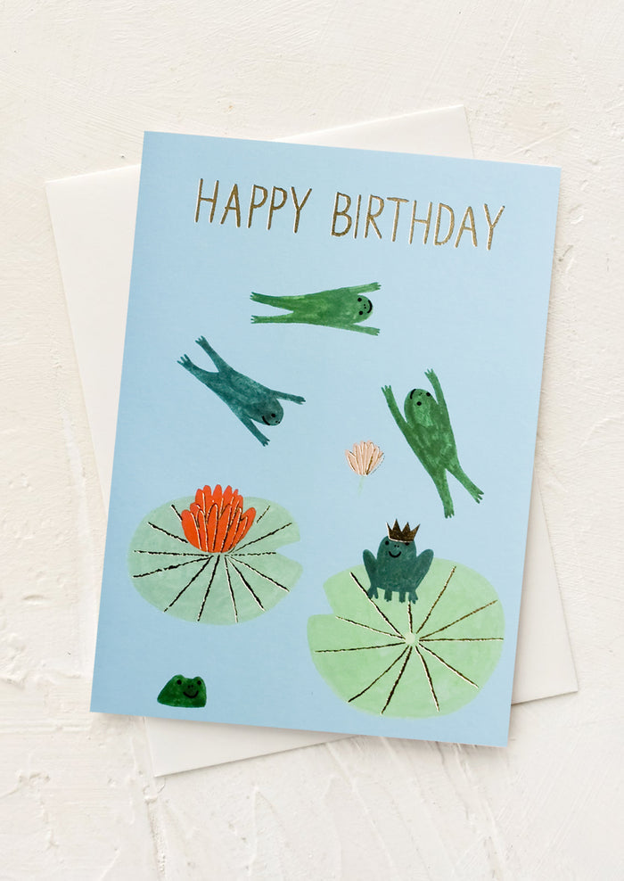 A greeting card with illustrated frogs jumping around on lilypads.