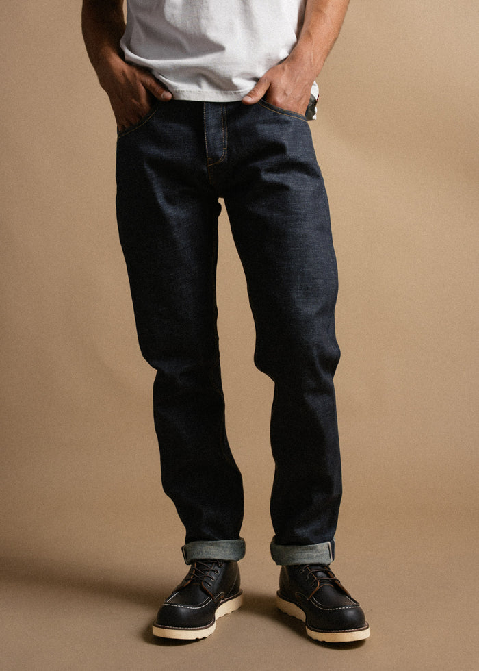 Man in dark jeans and boots stands casually with hands in pockets against a beige background, wearing a white shirt.