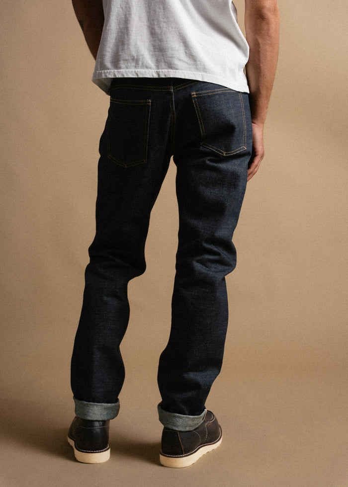 Man in dark jeans and white shirt stands against a beige background, showcasing rolled cuffs and brown shoes.