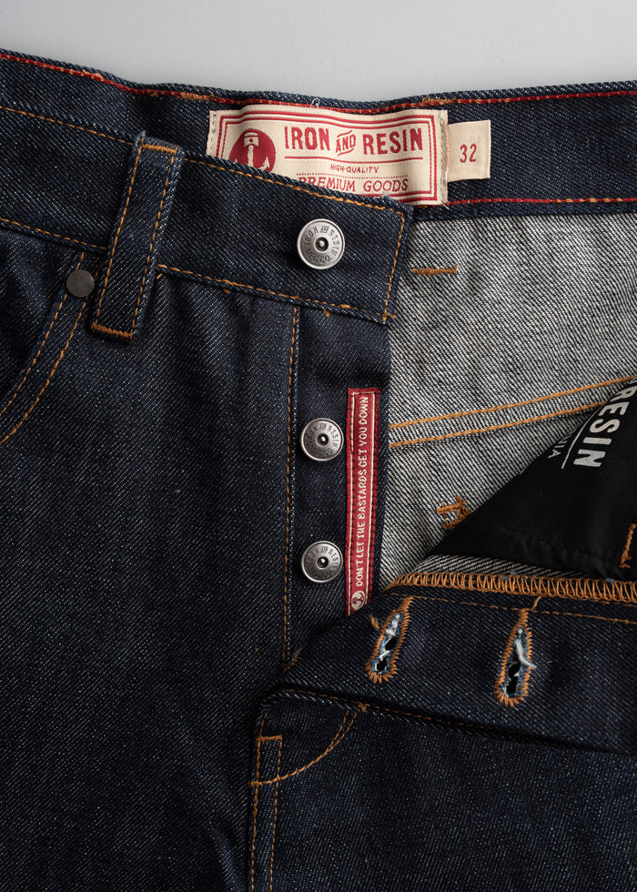 Close-up of Iron and Resin jeans with button fly, detailed stitching, and label showing size 32. Denim texture is visible.