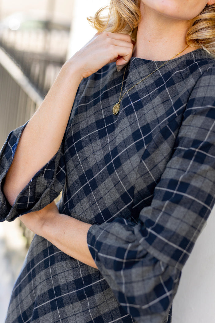 Close-up of a model wearing the Aspiga Elizabeth Check Dress in Blue/Grey, showing the neckline and necklace, hand near collar