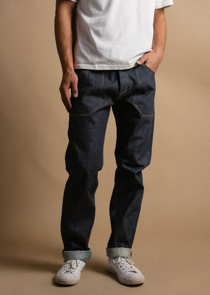 Man in white t-shirt and dark jeans stands casually, hands in pockets, wearing white sneakers against a neutral background.