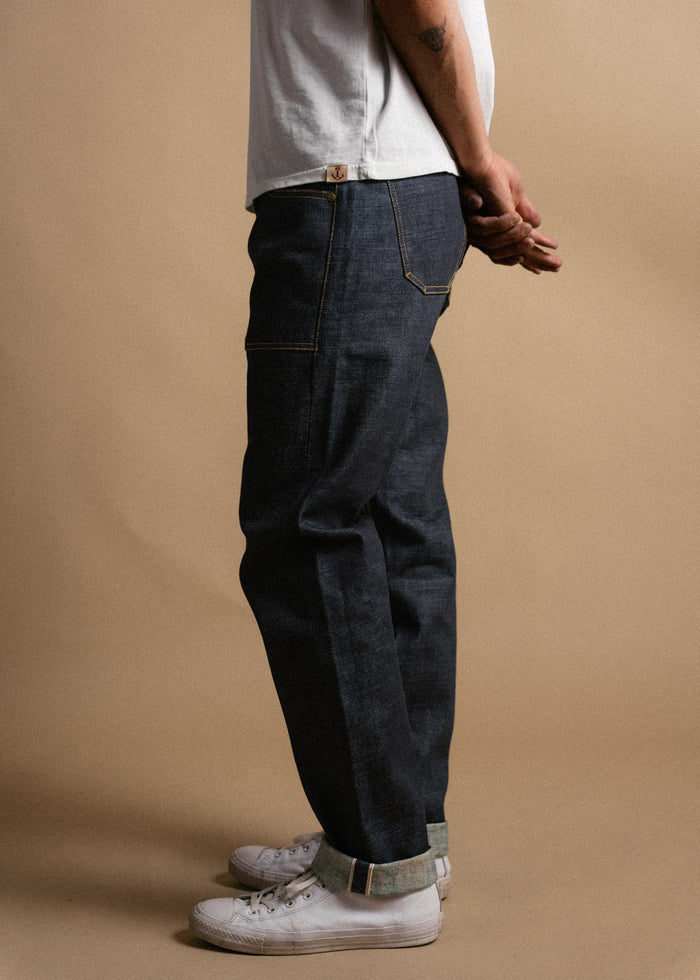 Man in white shirt and rolled-up jeans stands sideways, hands clasped. White sneakers on a beige background. Casual style.