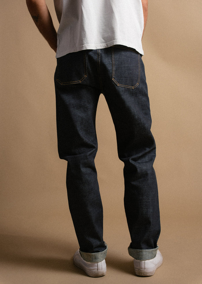 Person in dark denim jeans, white sneakers, and a white shirt stands against a beige background, facing away from the camera.
