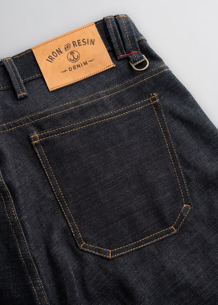 Dark denim jeans with a tan "Iron and Resin" label on the waistband, featuring contrast stitching and a metal ring detail.