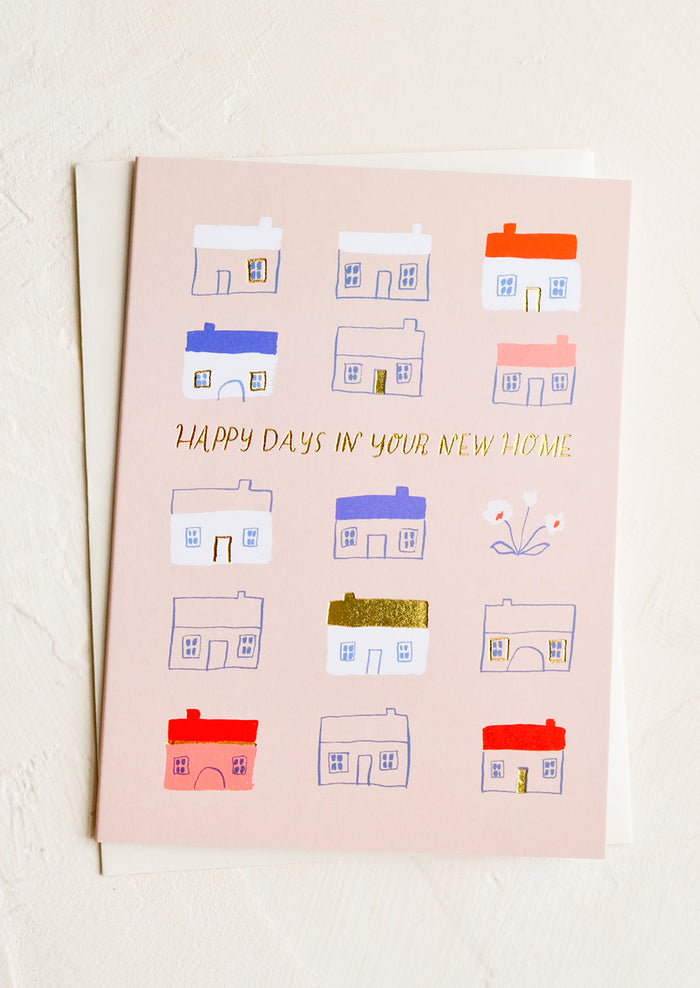 A pink greeting card with illustrated red and blue houses, text reads "Happy days in your new home".