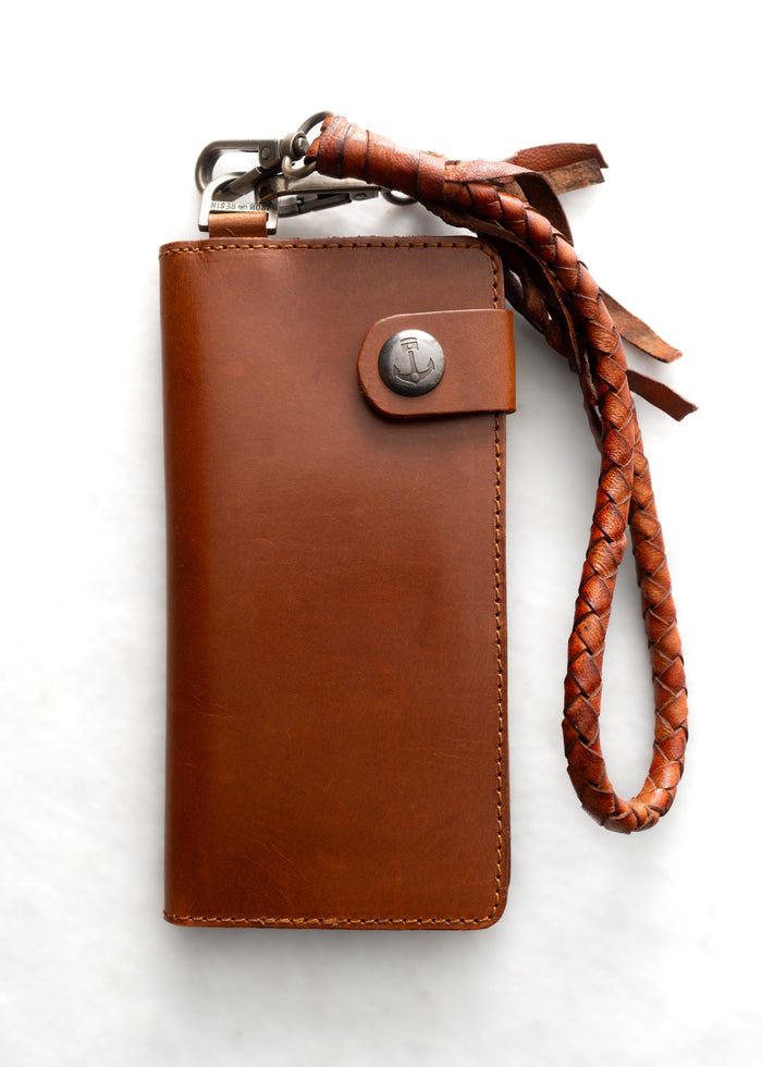 Brown leather wallet with braided strap and metal clasp on white background. Simple, elegant design with anchor emblem.