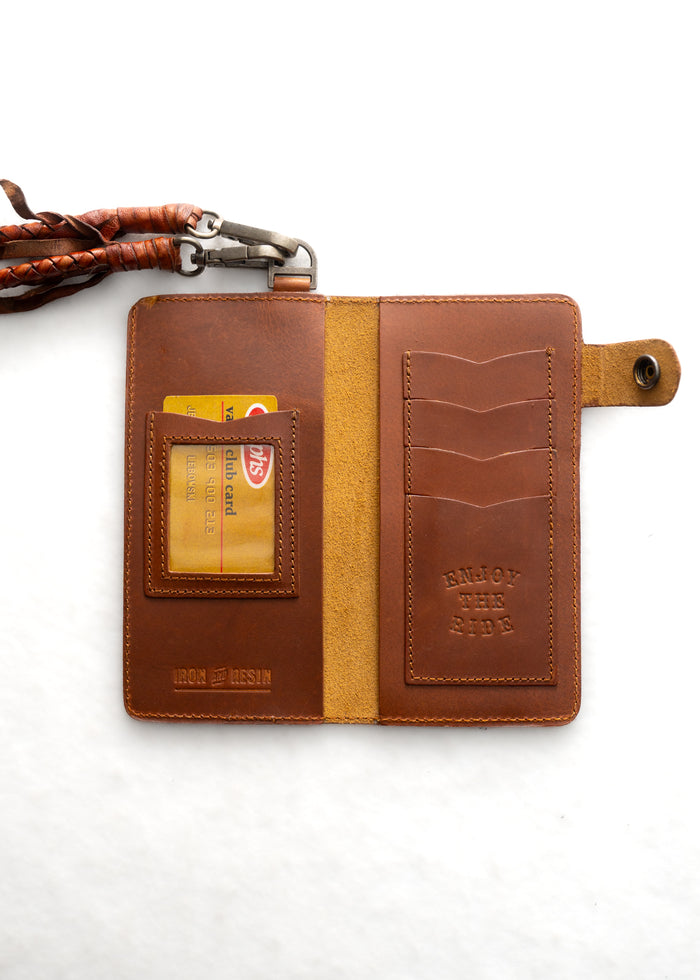 Open brown leather wallet with cards, embossed "Enjoy the Ride," attached braided strap, and metal clasp on white background.
