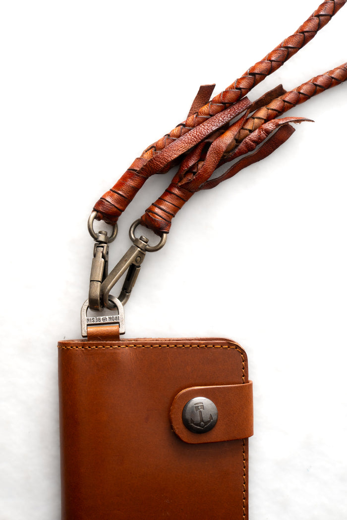 Brown leather wallet with braided straps and metal clasps on a white background. The wallet has a button with an anchor design.