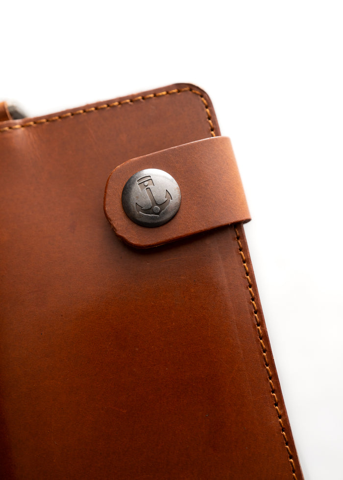 Brown leather notebook with stitched edges, featuring a metal anchor button closure, on a white background.
