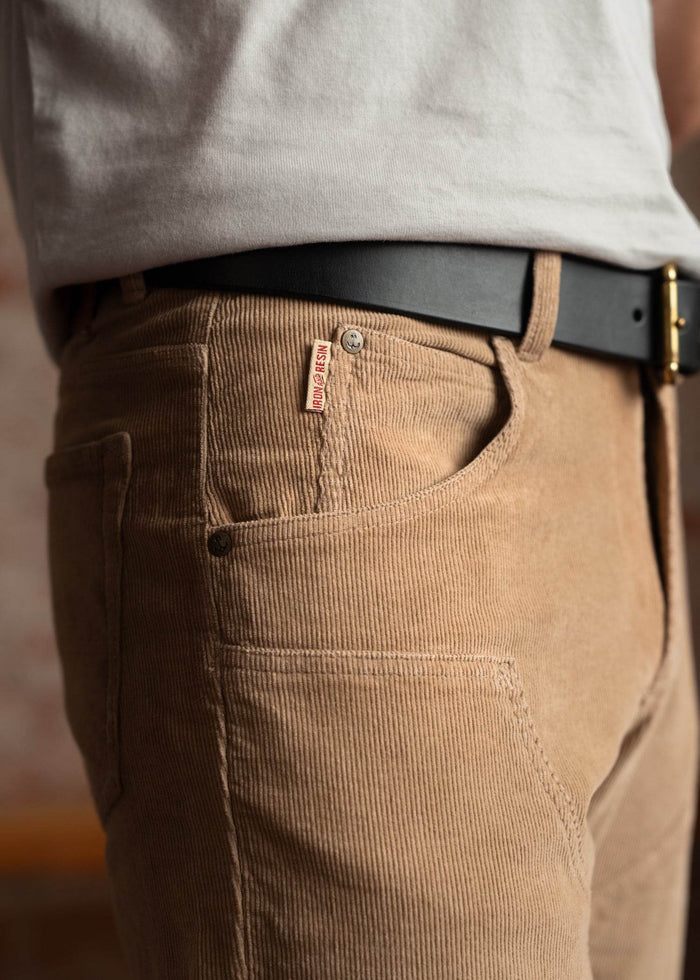 Close-up of a person wearing tan corduroy pants with a black belt and white shirt, showcasing the Iron and Resin label.