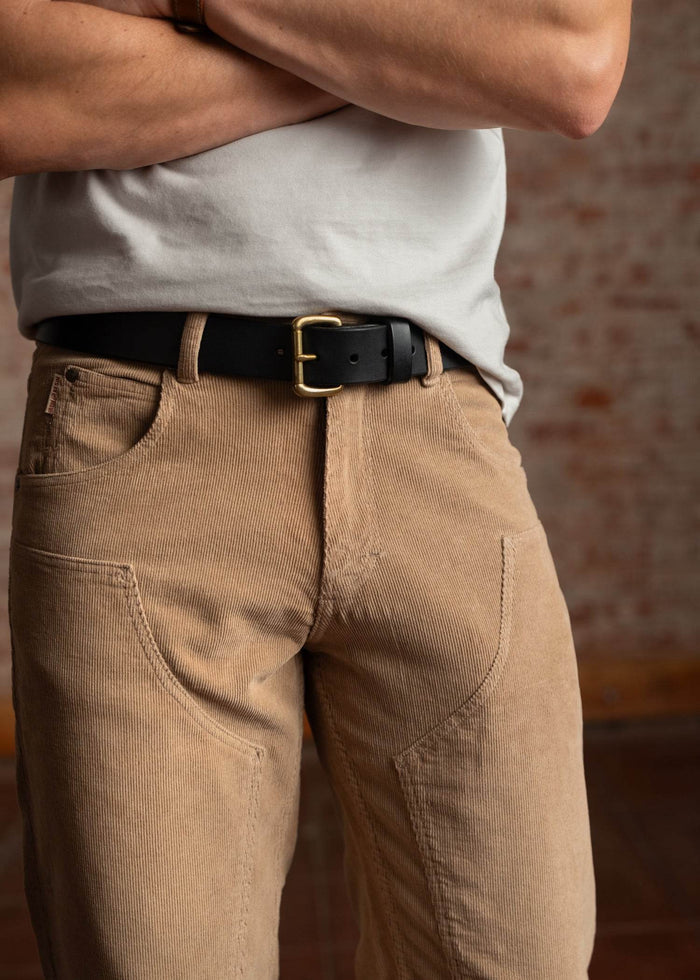 Man in a white shirt and tan corduroy pants with a black belt stands against a brick wall, arms crossed casually.