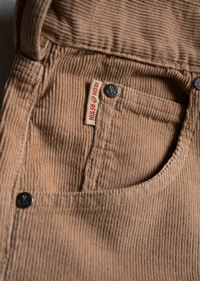 Close-up of tan corduroy pants pocket with Iron and Resin label and black buttons, highlighting texture and brand detail.