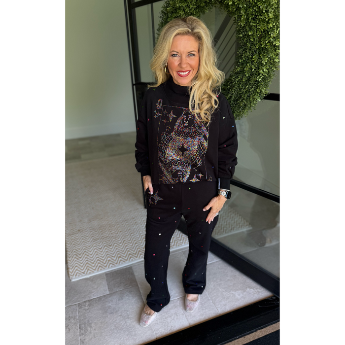 A woman stands indoors wearing a black matching lounge set with a large multicolor cat graphic on the sweatshirt and small star-like embellishments on the pants. The setting includes a glass door, tiled floor, and a green wreath in the background.