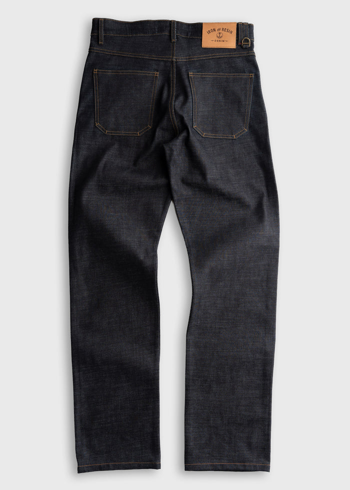 Dark denim jeans with a leather patch on the waistband, featuring two back pockets and contrast stitching, laid flat on a surface.