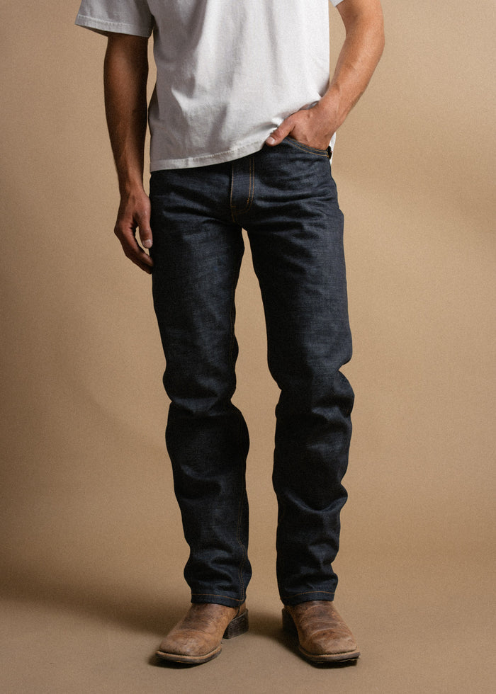 Man in white shirt and dark jeans stands against a beige background, hands in pockets, wearing brown boots. Casual and relaxed style.