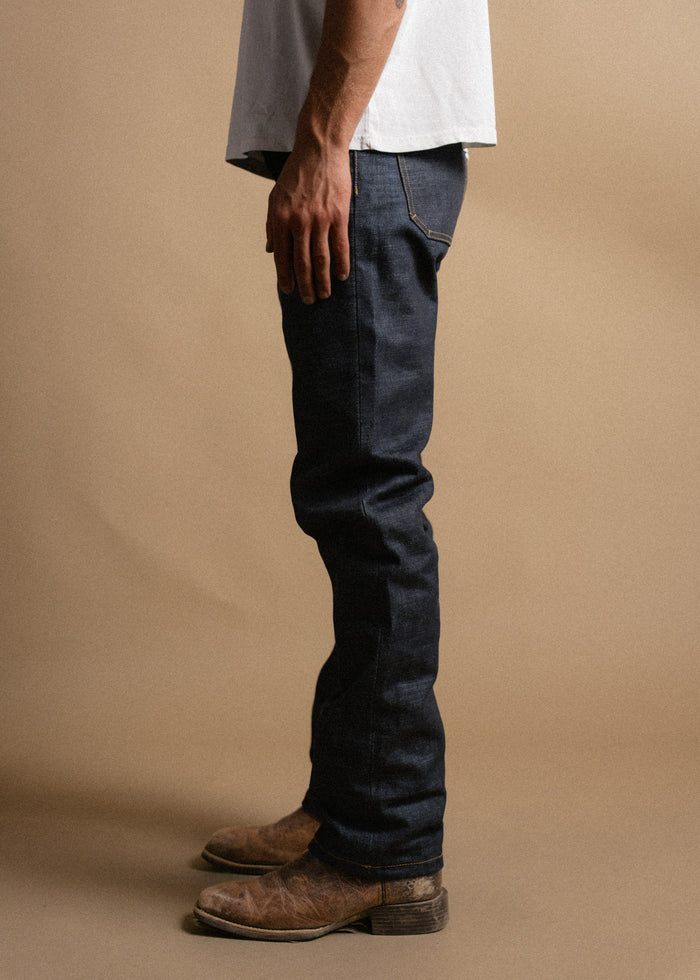 Man in a white shirt and dark jeans stands sideways, wearing worn brown boots, against a plain beige background.