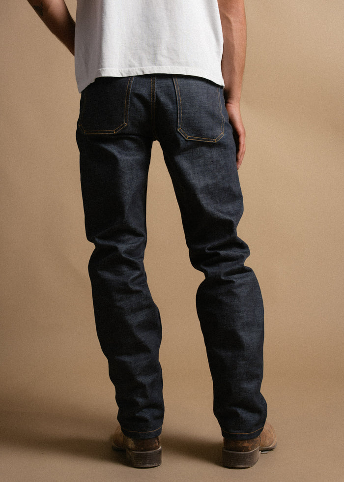Man in a white shirt and dark jeans stands facing away on a beige background, showcasing denim fit and style.