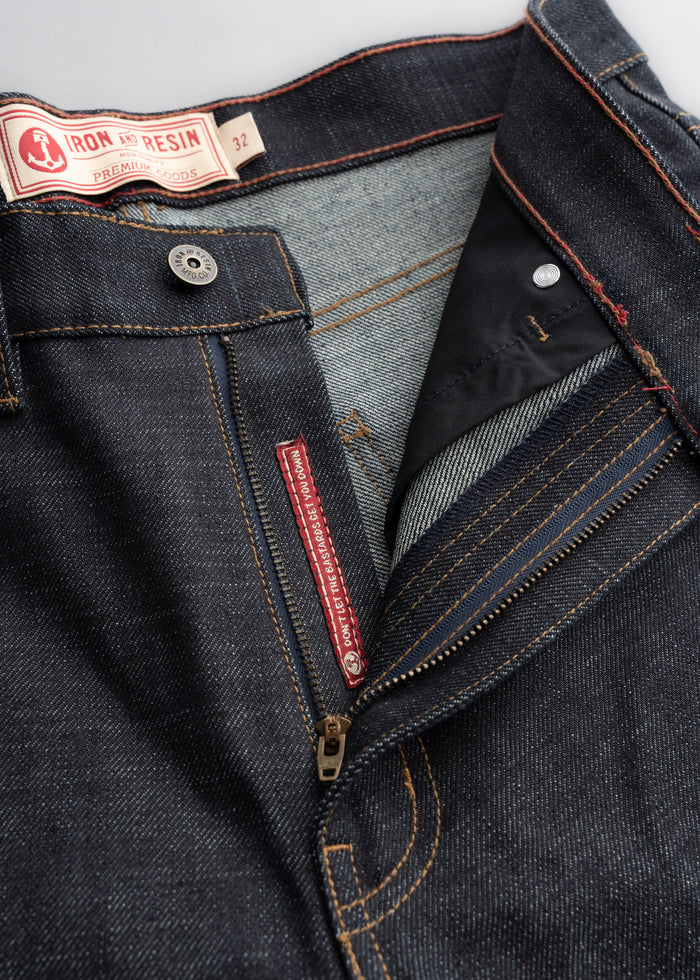 Close-up of Iron and Resin jeans with a visible zipper and label. Dark denim with orange stitching, showcasing premium quality.