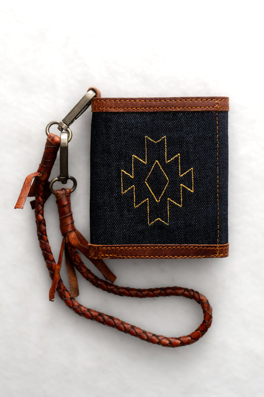 Denim wallet with brown leather trim and braided strap, featuring a yellow geometric pattern on a white background.