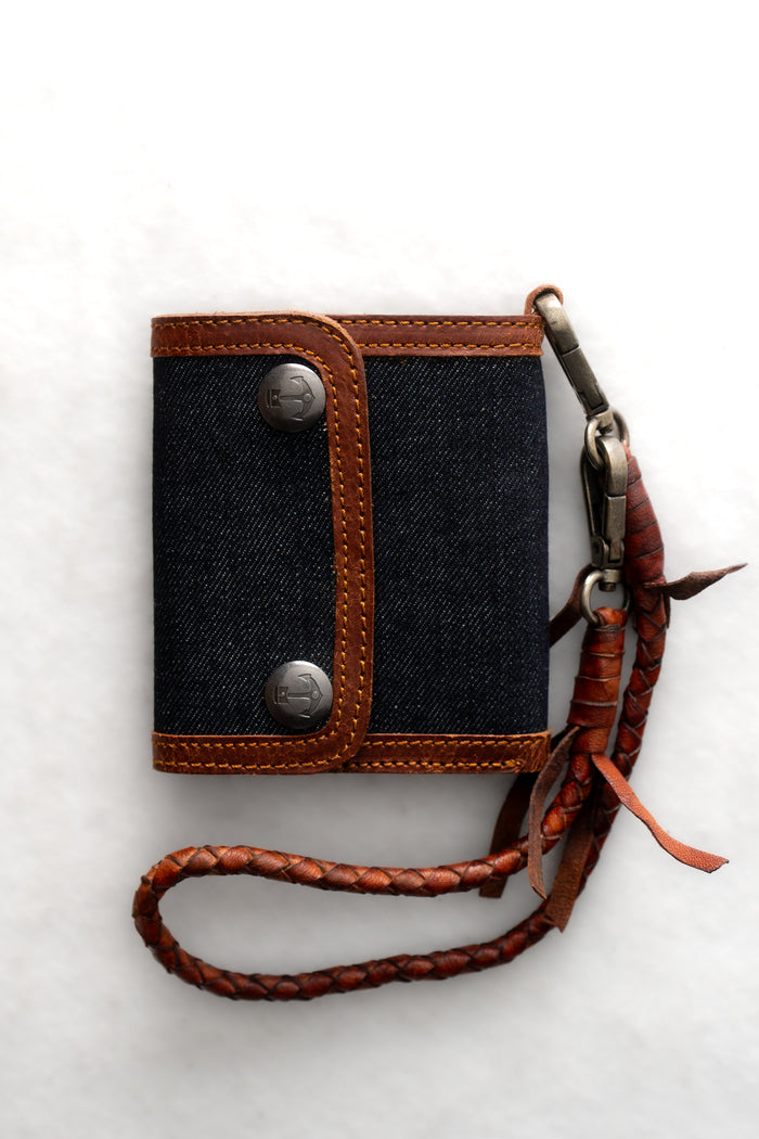 Denim wallet with brown leather trim and braided strap on a white background, featuring metal buttons for closure.