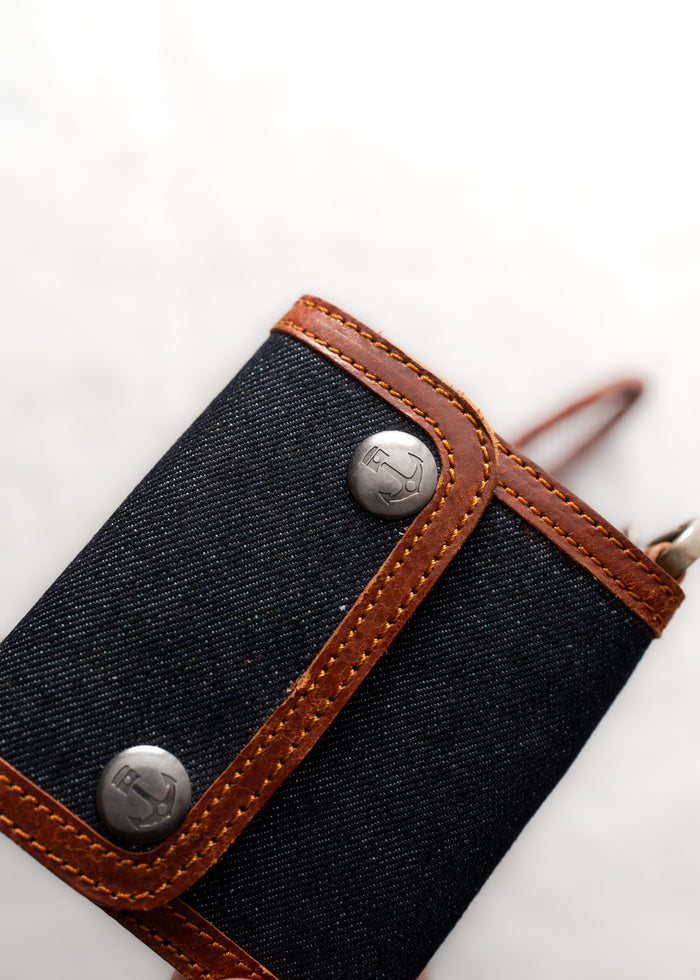 Close-up of a denim wallet with brown leather trim and metal buttons on a light background.