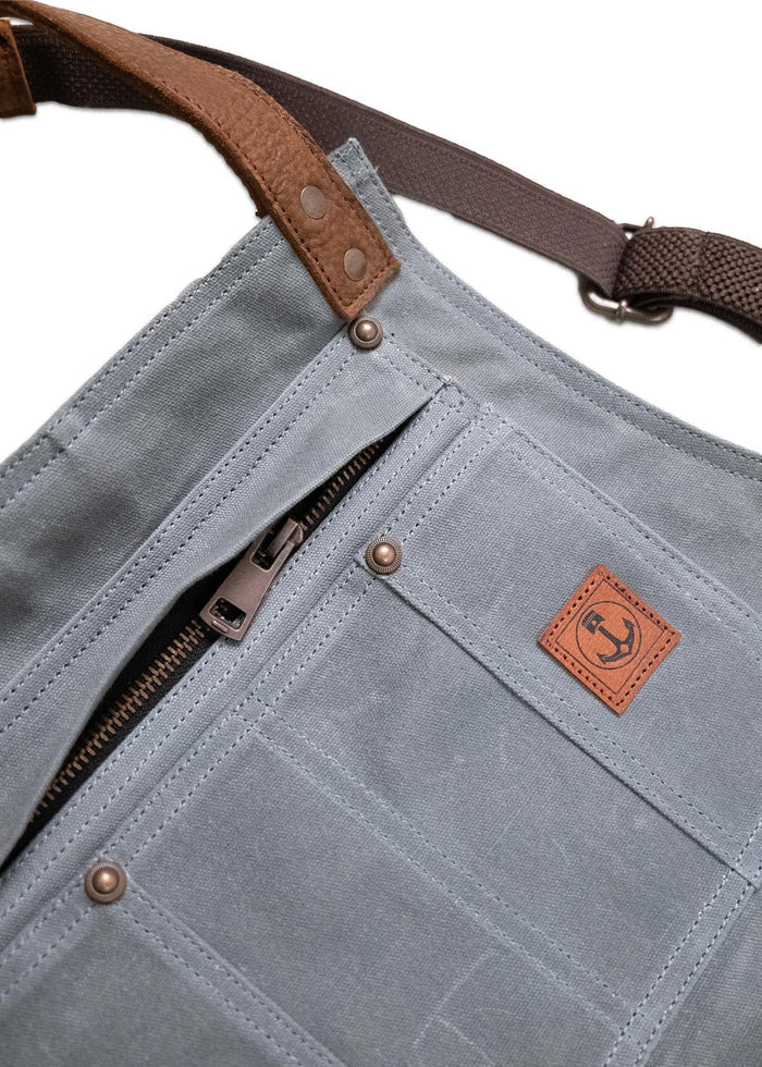 Blue canvas bag with brown leather strap, metal zipper, and anchor logo patch. Durable design with visible stitching and rivets.