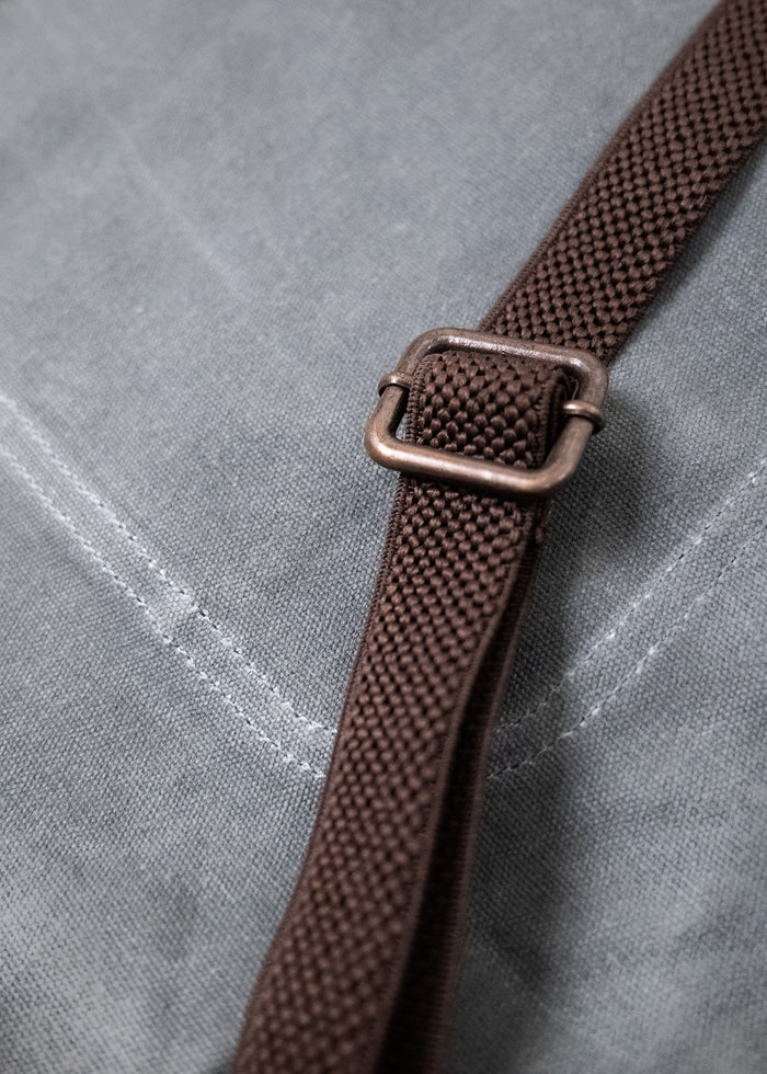 Close-up of a brown strap with a metal buckle on a gray fabric surface, showing texture and stitching detail.