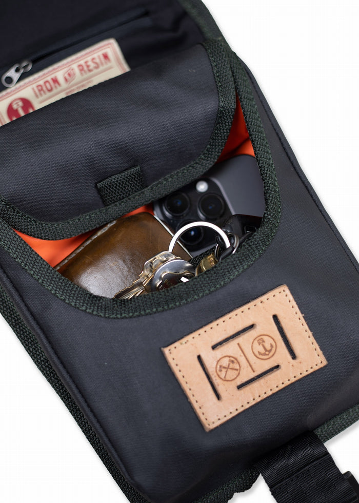 Open black bag with leather patch, showing a phone, keys, and wallet inside. Iron & Resin label visible in the pocket.