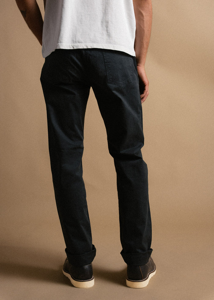 Man in black pants and white shirt stands on a beige background, showcasing casual style with rolled cuffs and brown shoes.