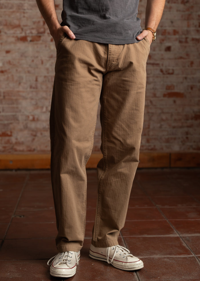 Man in khaki pants and white sneakers stands against a brick wall, hands in pockets, wearing a gray shirt. Casual style.