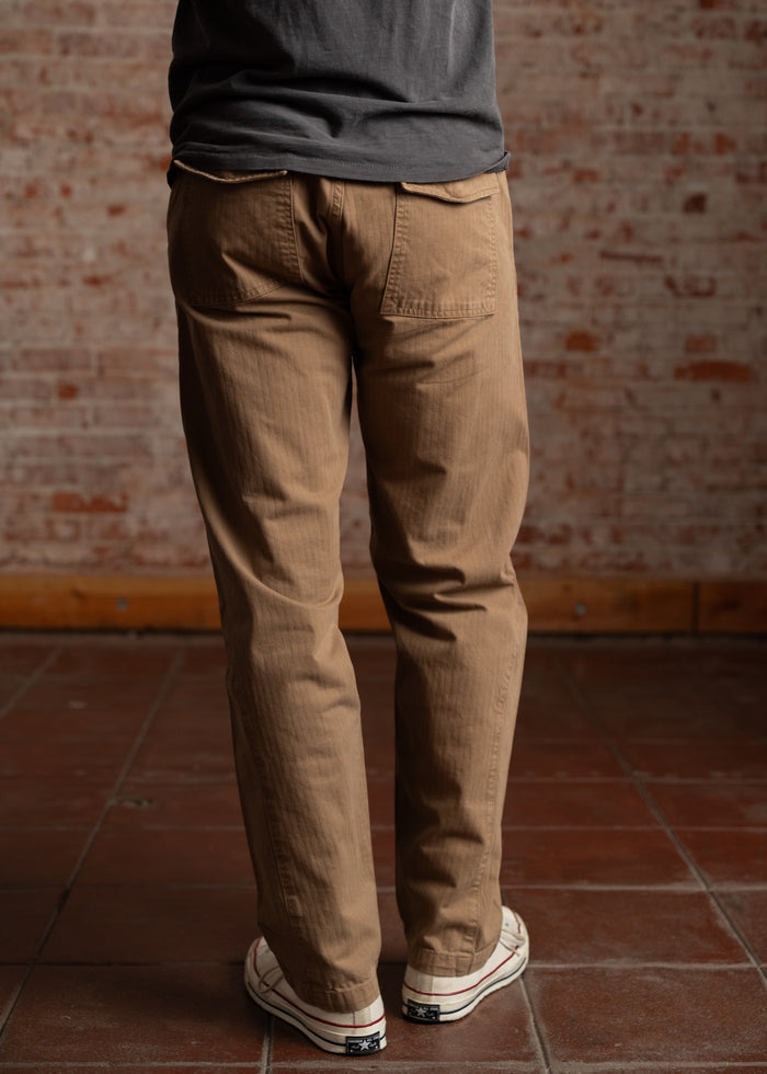 Person in khaki pants and sneakers stands on a tiled floor against a brick wall, with hands in pockets, facing away.