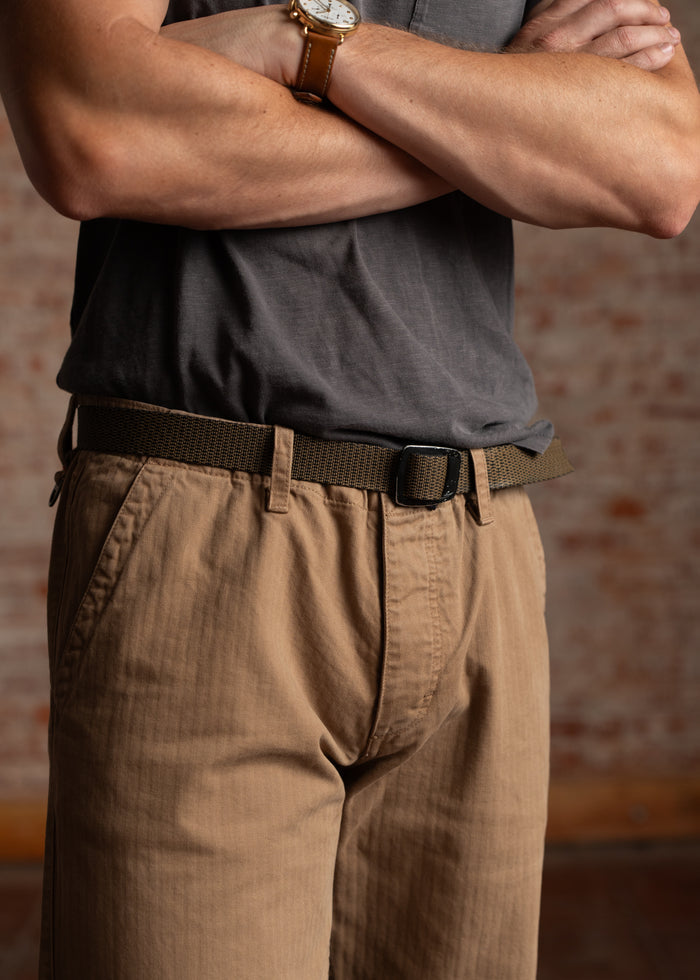 Man in a gray shirt and khaki pants, arms crossed, wearing a watch, stands against a brick wall.