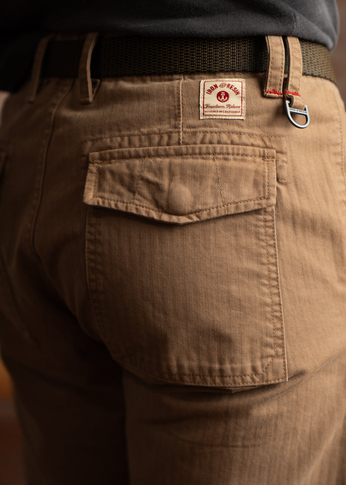 Close-up of brown pants with a back pocket, featuring an Iron and Resin label and a black belt looped through the waistband.